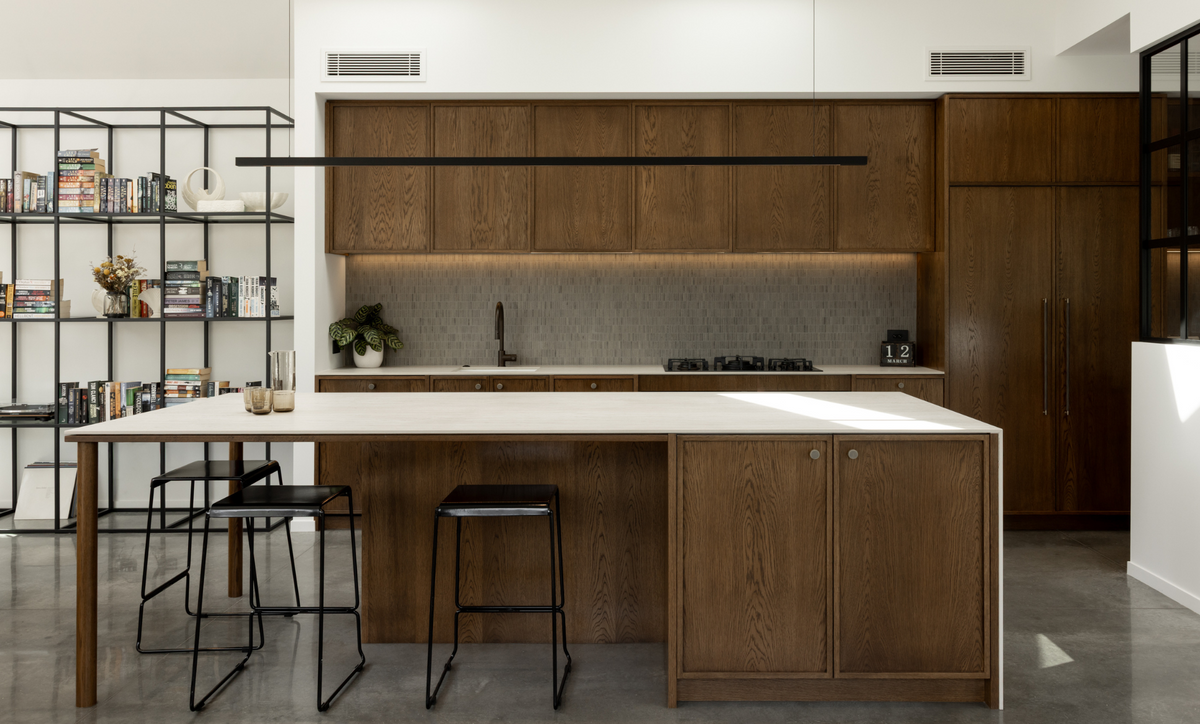 A modern kitchen island with medium brown wood cabinets and a white countertop, set in a bright contemporary kitchen with black bar stools, a black shelving unit, and skylights.