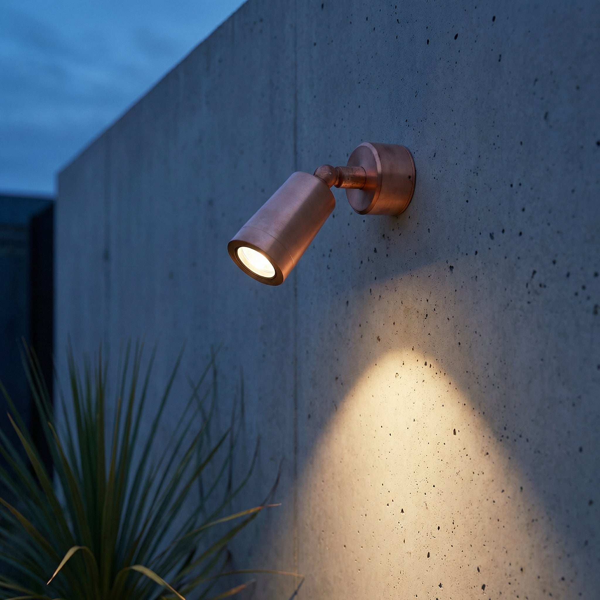 Copper outdoor wall light fixture on a concrete wall with a plant in the foreground.