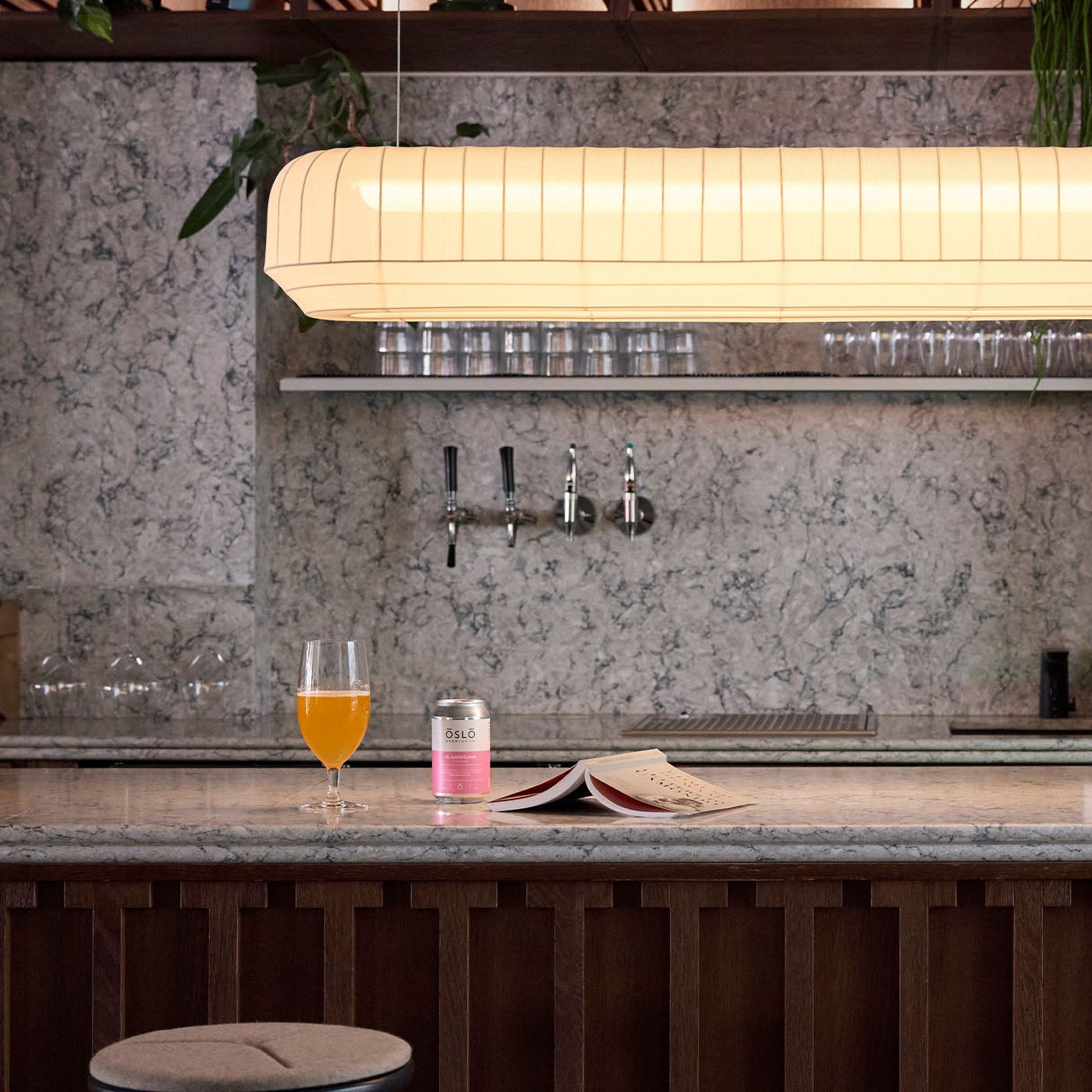 Bar counter with a glass of beer and a can, featuring a modern light fixture and marble backsplash.