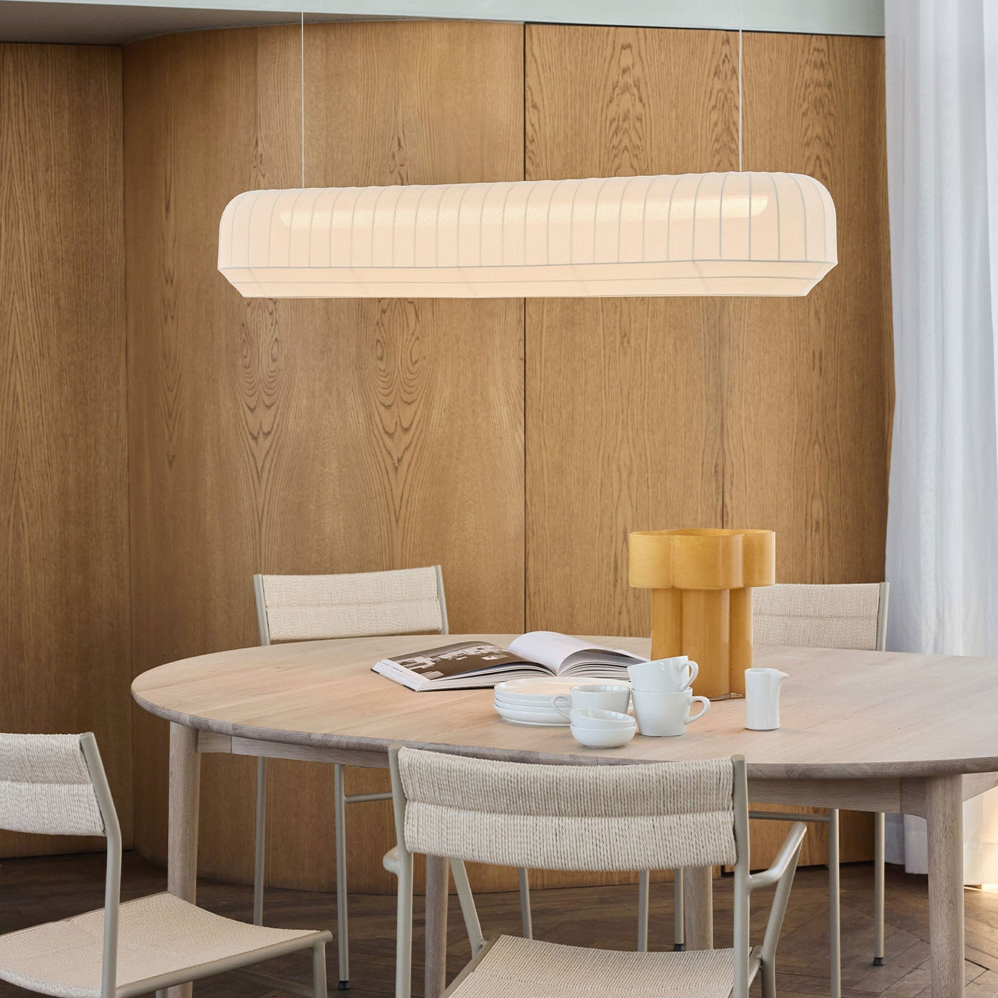 Dining area with a round table, chairs, and a linear pendant light against a wooden paneled wall.