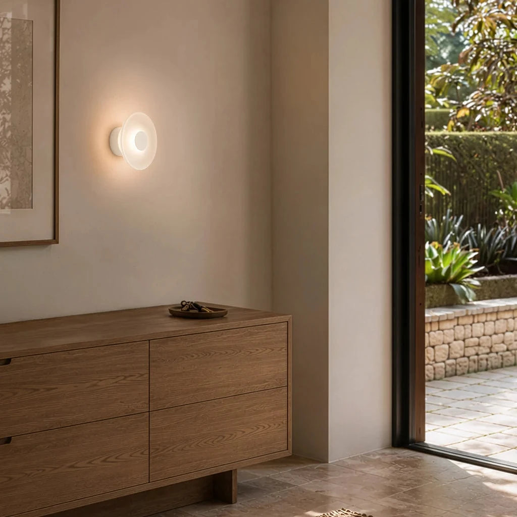 A modern wall-mounted light fixture with a circular white shade and warm glowing bulb, installed on a beige wall above a wooden credenza in a minimalist interior setting.