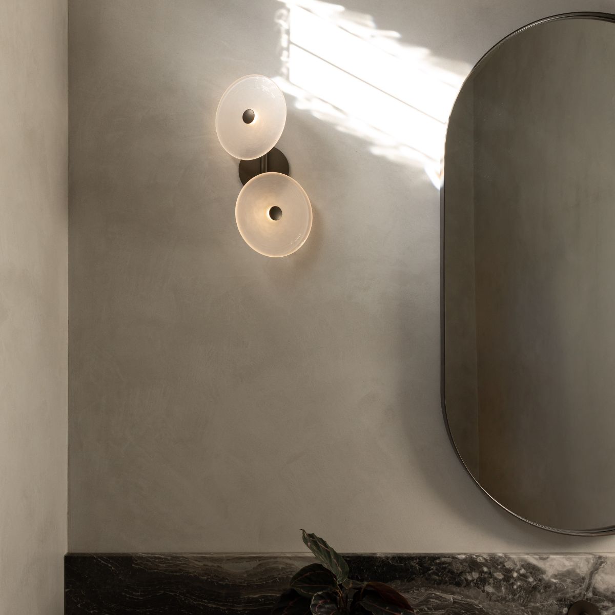 A modern wall-mounted double-light fixture with two frosted circular shades and a dark metal mount, installed on a textured beige plaster wall next to an oval mirror.
