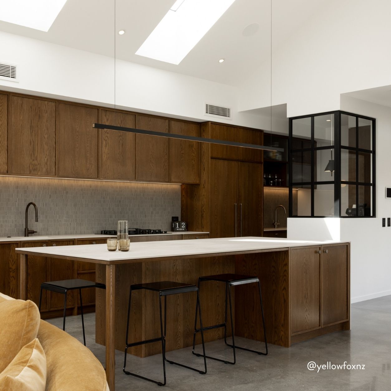 A modern kitchen island with a white countertop and dark wood cabinetry, styled in a bright contemporary kitchen with black bar stools and a long linear pendant light.