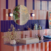 A vibrant bathroom interior featuring bold vertical striped wallpaper in shades of pink, purple, and orange, a matching vanity counter, a white rectangular sink, a round wall mirror, and decorative floral arrangements.