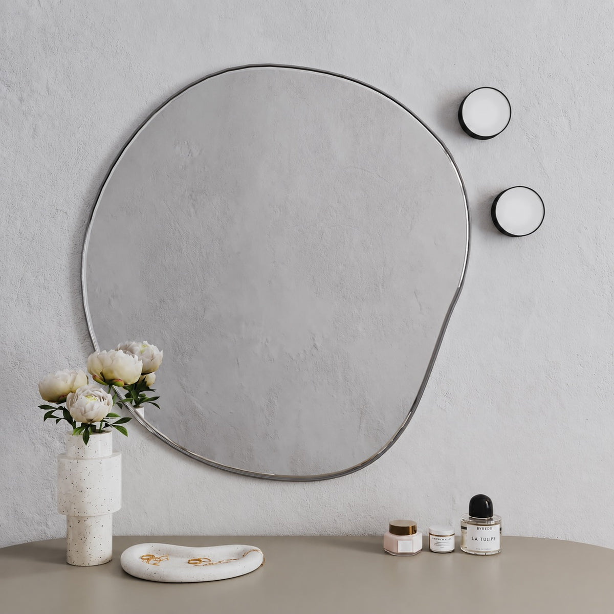 An organic-shaped wall mirror with a thin black frame hangs on a textured light gray wall above a beige tabletop. A white vase with pale flowers, a small white dish, and a few cosmetic jars are arranged below, with two matching round wall sconces visible on the right.